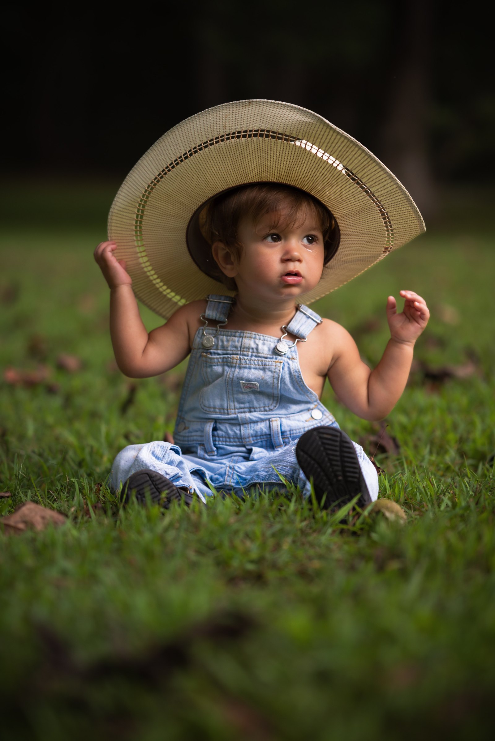 Young child in denim overalls sitting in the grass beneath an oversized cowboy hat