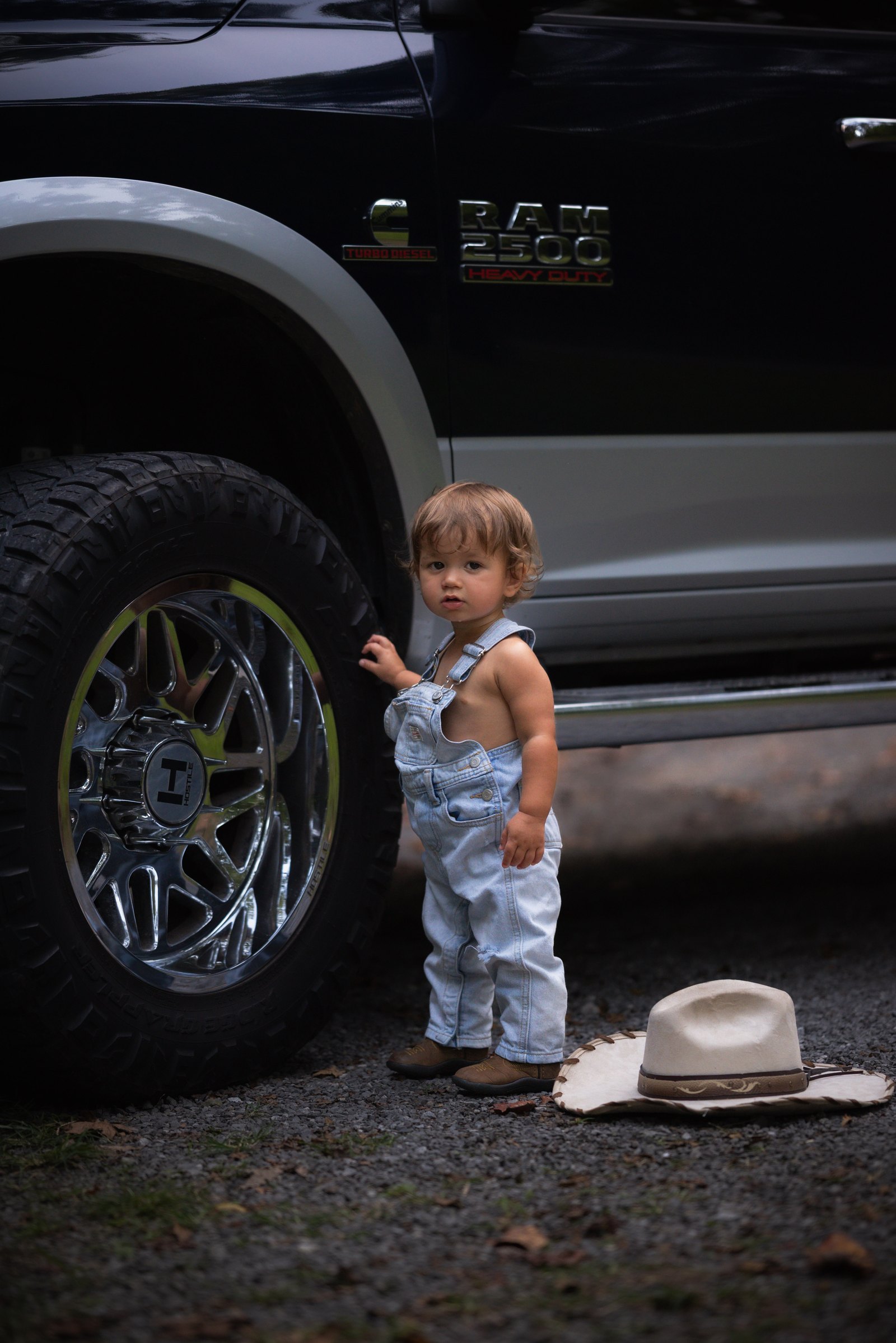 Young child in denim overalls standing beside a truck and cowboy hat