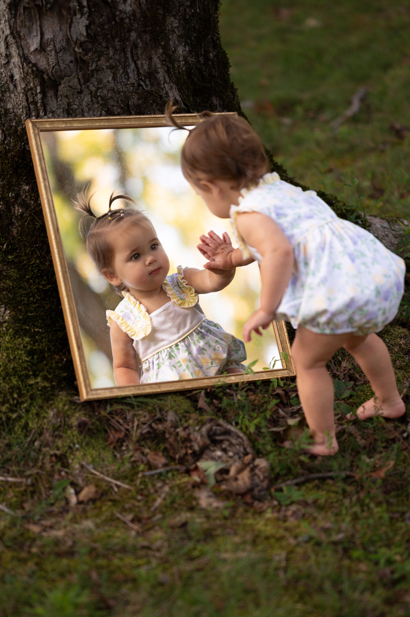 Young child reaching toward her reflection in a mirror beside a woodland tree